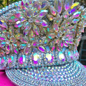 May include: A white hat with a large brim, covered in iridescent rhinestones and crystals. The hat is decorated with large, flower-shaped clusters of rhinestones.