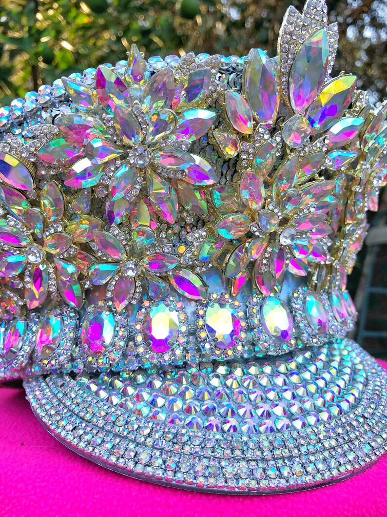 May include: A white hat with a large brim, covered in iridescent rhinestones and crystals. The hat is decorated with large, flower-shaped clusters of rhinestones.