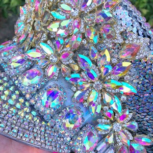 May include: A close-up of a silver and iridescent embellished hat with a variety of rhinestones and sequins. The hat is decorated with large iridescent flower-shaped embellishments.
