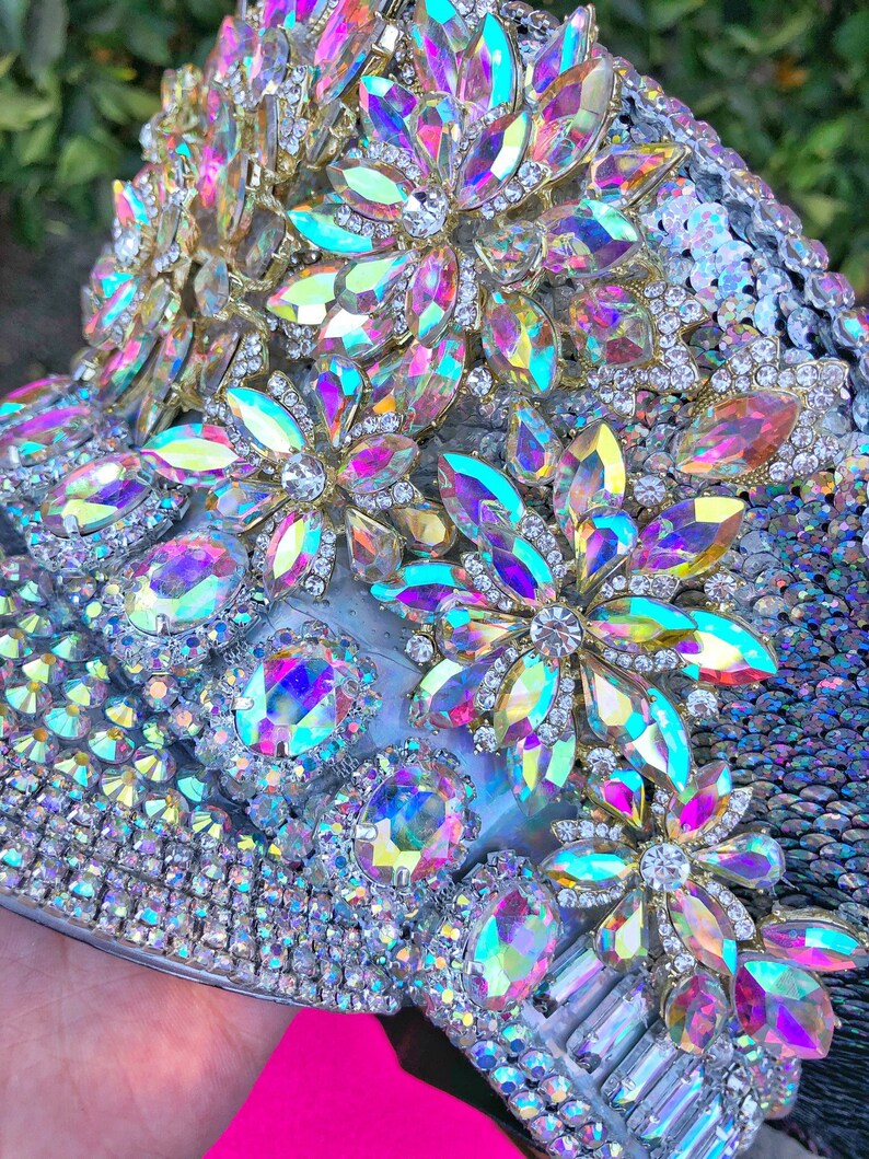 May include: A close-up of a silver and iridescent embellished hat with a variety of rhinestones and sequins. The hat is decorated with large iridescent flower-shaped embellishments.