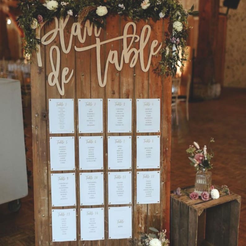 Peut inclure: Un panneau en bois rustique avec les mots "Plan de Table" écrits en cursive. Le panneau est décoré de verdure et de fleurs. Il y a 14 cartes blanches attachées au panneau, chacune avec un numéro de table et une liste de noms.