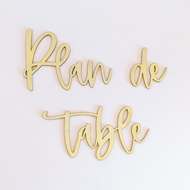 May include: Wooden sign with the words "Plan de Table" in a cursive font. The sign is painted in a light wood color.