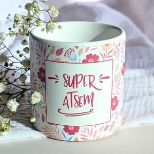 Peut inclure: Mug en céramique blanche avec un motif floral rose, bleu et pêche. Le mug porte l'inscription "SUPER ATSEM" en rose dans un cadre rectangulaire. Le motif comprend des feuilles et des fleurs.