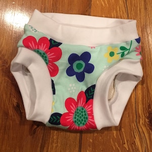 May include: White training pants with a light blue background and a floral print featuring pink, red, blue, green, and yellow flowers. The pants have a white elastic waistband.