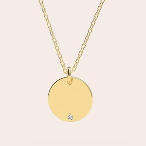 May include: A gold-colored necklace featuring a delicate chain and a circular pendant. The pendant has a small, sparkling gemstone near the bottom. The necklace is displayed against a soft, neutral background.