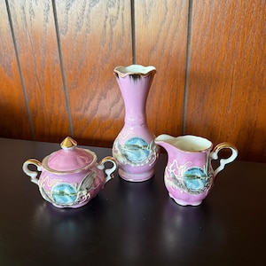 May include: A pink ceramic tea set with gold trim, featuring a sugar bowl with a lid, a creamer, and a vase. Each piece has a scenic image of Niagara Falls. The set is displayed on a dark surface against a wood background.