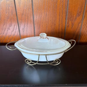 May include: Oval white casserole dish with a clear glass lid and a decorative gold-colored metal stand. The lid has a clear glass knob. The dish has gold-colored handles on each side.