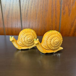 May include: Two ceramic snail figurines with yellow and orange tones. The snail shells have a spiral design, and the word "FLORIDA" is printed on the base. The figurines are on a dark brown surface.