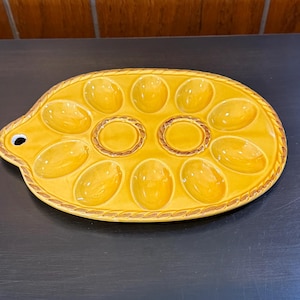 May include: Yellow ceramic egg serving platter with 12 oval indentations and two round ones. The platter has a decorative rope-like border and a small handle with a hole. The serving dish is on a dark surface.