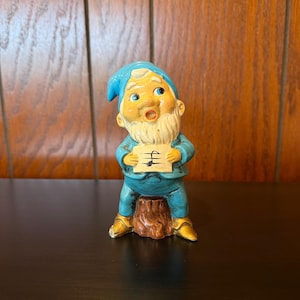 May include: A ceramic garden gnome figurine, wearing a blue hat and outfit, with a white beard and holding a sign with musical notes. The gnome is sitting on a brown tree stump, with gold-colored shoes.