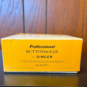 May include: A yellow box with the text "Professional Buttonholer by SINGER for Slant Needle Zig-Zag Sewing Machines Part No. 102577" and "A Trademark of THE SINGER COMPANY Printed in Great Britain".