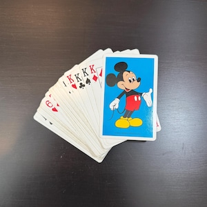 May include: A deck of playing cards with a Mickey Mouse card on top. The Mickey Mouse card has a blue background and features Mickey in his classic red shorts, yellow shoes, and white gloves. The other cards show the suits of hearts, diamonds, clubs, and spades.