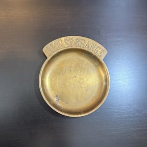 May include: A round, brass pocket change dish with the words "POCKET CHANGE" arched across the top. The dish is a warm gold color and has a slightly worn appearance.