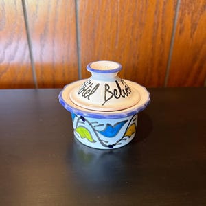 May include: A small, ceramic container with a lid. The container is white with blue, yellow, and green painted designs. The lid has the words "Hôtel Belie" written on it in black script. The rim of the lid and the top of the container are trimmed in blue.