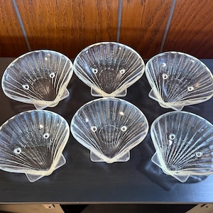 May include: A set of six clear glass dishes shaped like scallop shells. Each dish features a ribbed texture radiating from the base to the scalloped edge and includes three small feet on the underside for stability when placed on a flat surface.