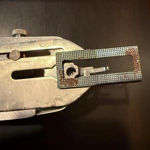 May include: Close-up of a silver sewing machine foot with a rectangular metal plate. The plate has a small opening and a textured surface with a rusty patina.
