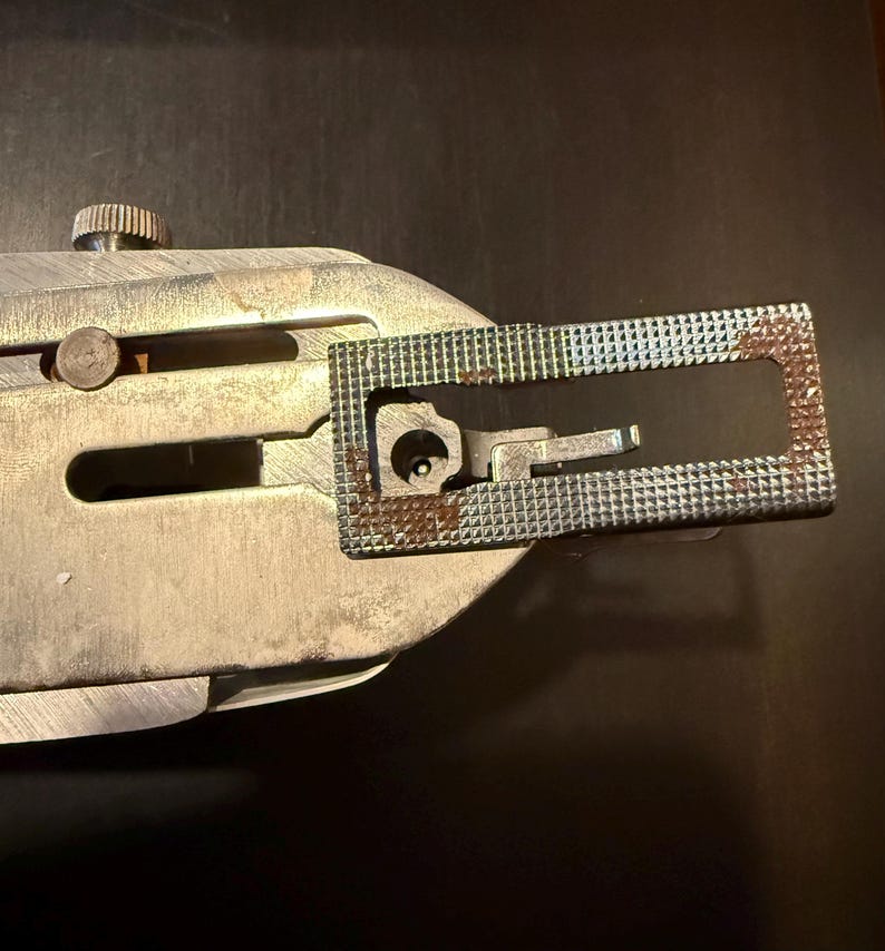 May include: Close-up of a silver sewing machine foot with a rectangular metal plate. The plate has a small opening and a textured surface with a rusty patina.