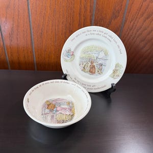 May include: A white ceramic plate and bowl set with illustrations and text. The plate features a scene with a girl and a house, with text around the rim. The bowl has a similar illustration and text. A charming vintage set.
