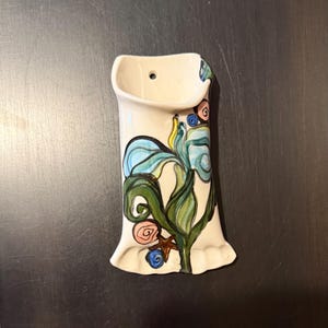 May include: A cream-colored ceramic wall pocket vase with a floral design. The vase features a blue iris with green leaves and pink and blue accents. The vase has a small hole at the top for hanging.