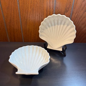 May include: Two white ceramic shell-shaped dishes. One is on a black stand, the other is on a dark surface. The dishes have a ribbed design, resembling a seashell. The background is a wooden wall.