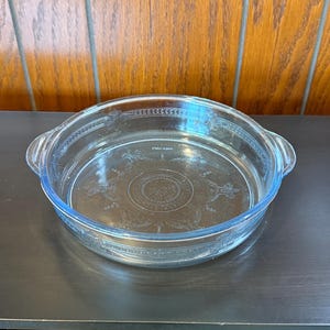 May include: Clear glass casserole dish with a round shape and two handles. The dish has a decorative floral pattern etched into the glass. The dish is sitting on a dark surface.