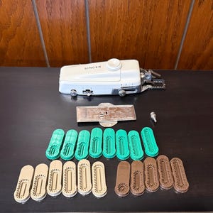 May include: A white Singer sewing machine with a green and brown plastic foot attachment. The machine is on a dark surface with a metal plate and 15 plastic feet.