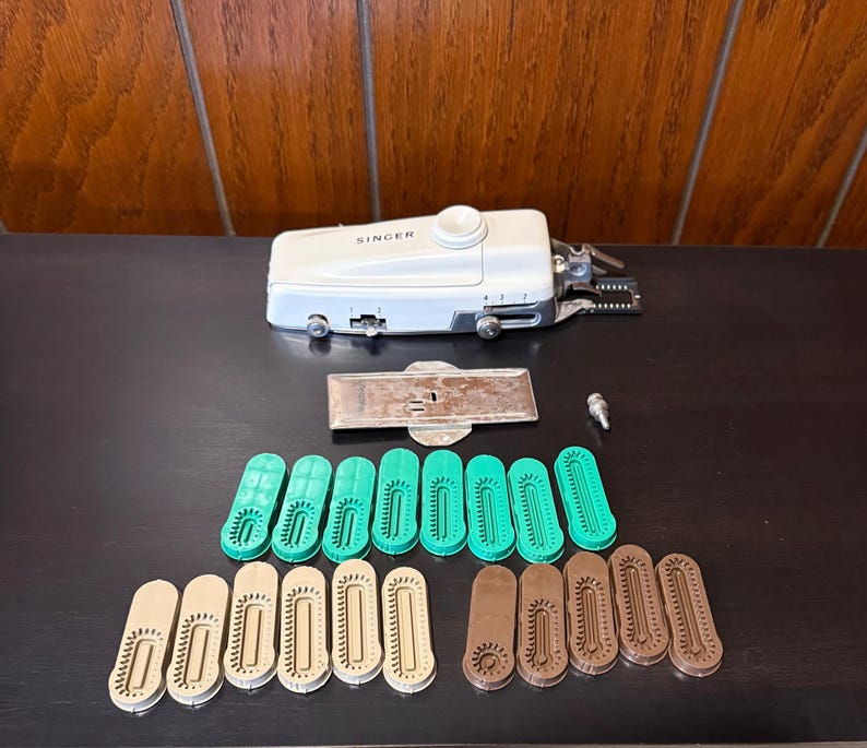 May include: A white Singer sewing machine with a green and brown plastic foot attachment. The machine is on a dark surface with a metal plate and 15 plastic feet.
