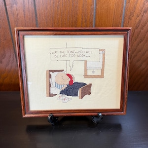 May include: Cross-stitch artwork in a wooden frame. The image depicts a person in bed, talking on the phone, with the text "...AT THE TONE...YOU WILL BE LATE FOR WORK..." above. The artwork is on a cream-colored background.