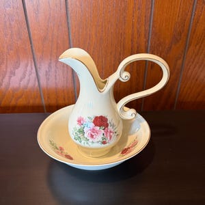 May include: Cream-colored vintage pitcher and basin set. The pitcher has a curved handle and a spout, with a floral design featuring red and pink roses. The basin has a matching floral pattern and gold trim. A classic decorative set.