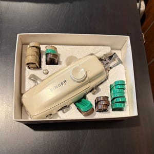 May include: A beige Singer brand sewing machine with a white knob and a green and brown plastic bobbin case. The machine is in a white box with a foam insert.