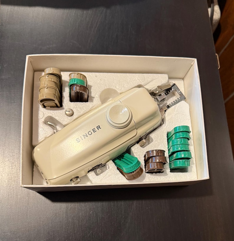 May include: A beige Singer brand sewing machine with a white knob and a green and brown plastic bobbin case. The machine is in a white box with a foam insert.