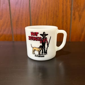 May include: White ceramic mug with a handle, featuring a graphic of a person with a dog and the text "HOT SPRINGS Arkansas" in red and black. The mug has a glossy finish and a curved handle.