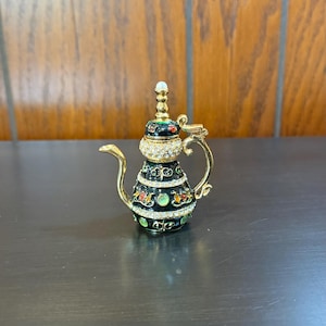 May include: A decorative black and gold teapot with a pearl-topped lid. The teapot is adorned with rhinestones and colorful floral accents. The handle and spout are gold-colored, adding to its ornate design. A unique collectible item.