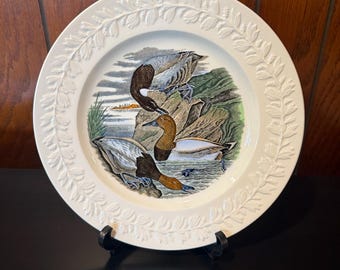 May include: A white decorative plate with a detailed illustration of ducks in a natural setting. The plate has a raised floral border and is displayed on a black stand. The ducks are depicted with brown, gray, and white feathers.