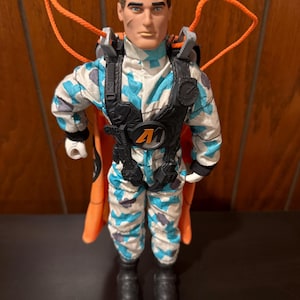 May include: Action figure wearing a blue and white camouflage jumpsuit with a black harness and orange parachute. The figure has black boots and a black helmet.