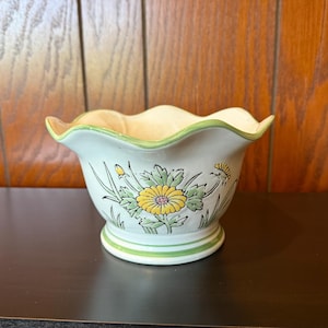 May include: A ceramic flower pot with a scalloped rim and a floral design. The pot is primarily white with a green rim and base. Yellow and green flowers are painted on the side of the pot.