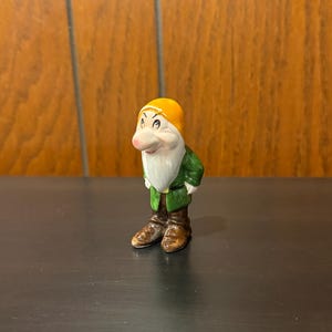 May include: A ceramic figurine of a dwarf from Snow White. The dwarf wears a yellow hat, green jacket, brown boots, and has a white beard. The figurine is standing on a dark surface.