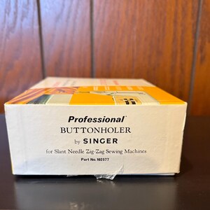May include: A white box with the text "Professional Buttonholer by SINGER for Slant Needle Zig-Zag Sewing Machines Part No. 102577"