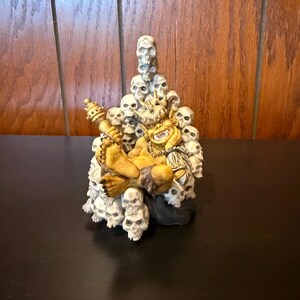 May include: A small, yellow, demonic figure sits on a throne made of white skulls. The figure has a crown, a club, and a fierce expression. The throne is surrounded by skulls.
