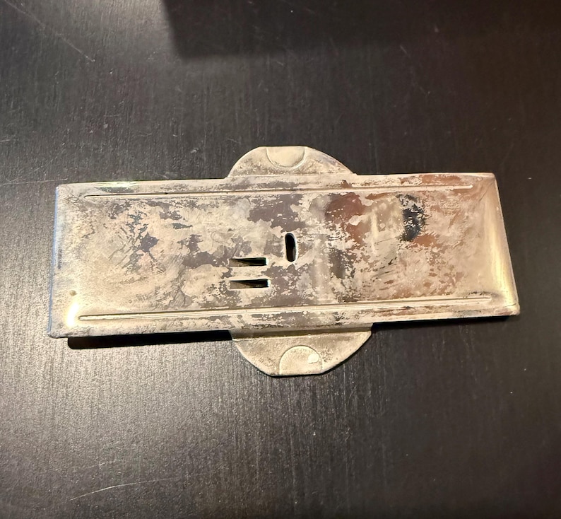 May include: A silver-toned metal plate with a keyhole and two small slots. The plate has a slightly worn, aged appearance.