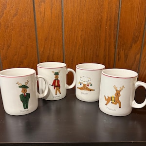 May include: Four white ceramic mugs with red trim and reindeer illustrations. Each mug has a different reindeer name: Donner, Dancer, Blitzen, and Prancer.