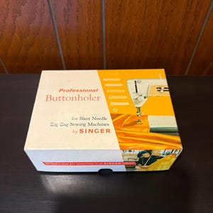 May include: A white box with a yellow and orange design. The box is labeled "Professional Buttonholer for Slant Needle Zig-Zag Sewing Machines by SINGER". The box features a vintage Singer sewing machine and a woman sewing.