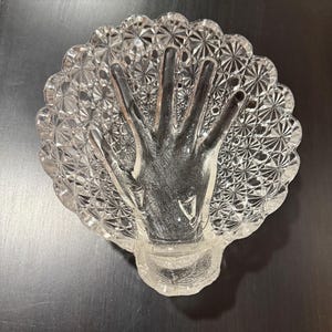 May include: A clear glass dish shaped like a scallop shell featuring a raised, molded hand design in the center. The surface of the dish is decorated with a repeating starburst pattern, and the edges are scalloped with a textured, faceted finish.