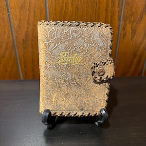 May include: A brown leather wallet with a textured surface and a braided black border. The wallet has a snap closure and the word "Bridge" in gold lettering. The wallet is displayed on a black stand.