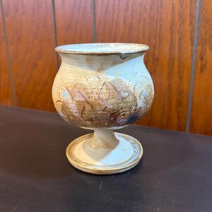 May include: A handmade ceramic goblet with a wide bowl and a pedestal base. The goblet features a beige and brown color scheme with abstract patterns. The rim is a lighter shade of beige. The goblet is displayed on a dark surface.
