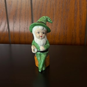 May include: A ceramic gnome figurine with a green hat and outfit, white beard, and rosy cheeks. The gnome is seated on a small, woven-looking stool. The figurine has a whimsical, vintage aesthetic.