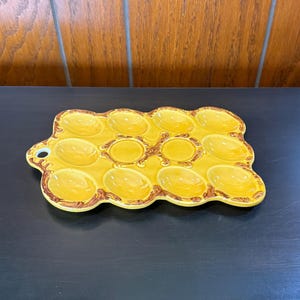 May include: A yellow ceramic egg serving tray with 12 oval indentations. The tray has a decorative brown border and a handle with a hole. The tray is designed to hold eggs for serving.