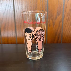 May include: Clear glass with a cartoon graphic of two figures and the words "love is" in red. The figures are nude with black hair and pink skin. The bottom of the glass reads "THE GREATEST THING MAN CAN FEEL".