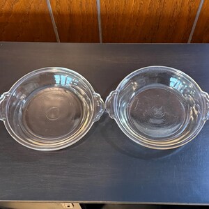 May include: Two clear glass pie dishes with scalloped handles. The dishes are round with a slightly raised rim and a flat bottom. They are sitting on a dark surface.