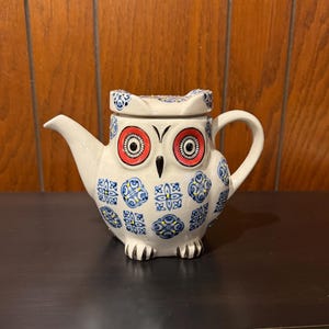 May include: White ceramic owl-shaped teapot with a lid. The teapot features large red and black eyes, a beak, and blue and yellow patterned designs. The spout and handle are white.